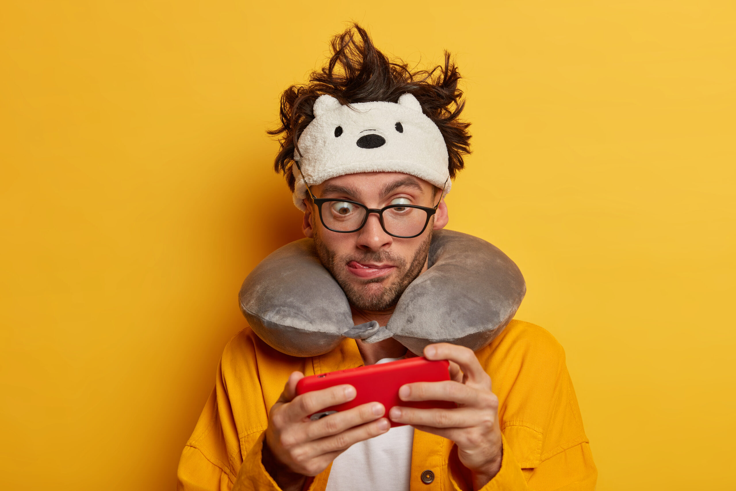 Addicted male passenger plays games online during trip, holds smartphone horizontally uses free wifi connection in plane wears sleeping attributes for transport, isolated on yellow waits for departure